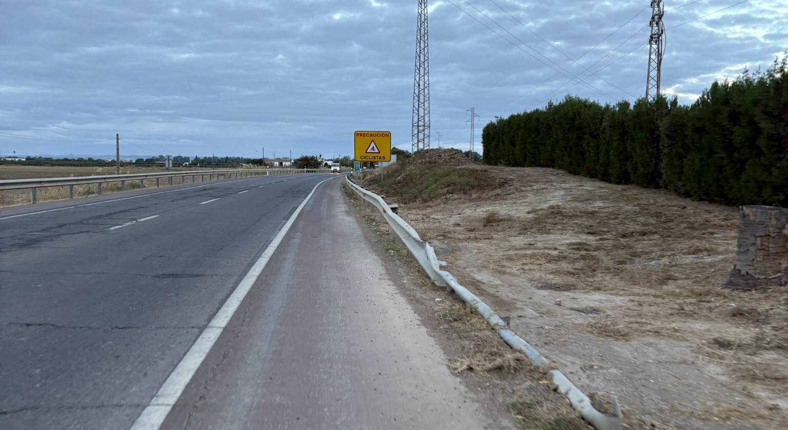 A wide two-lane Spanish road with a generous shoulder, stretching into the distance under clear skies. The surrounding landscape is relatively flat with sparse vegetation.