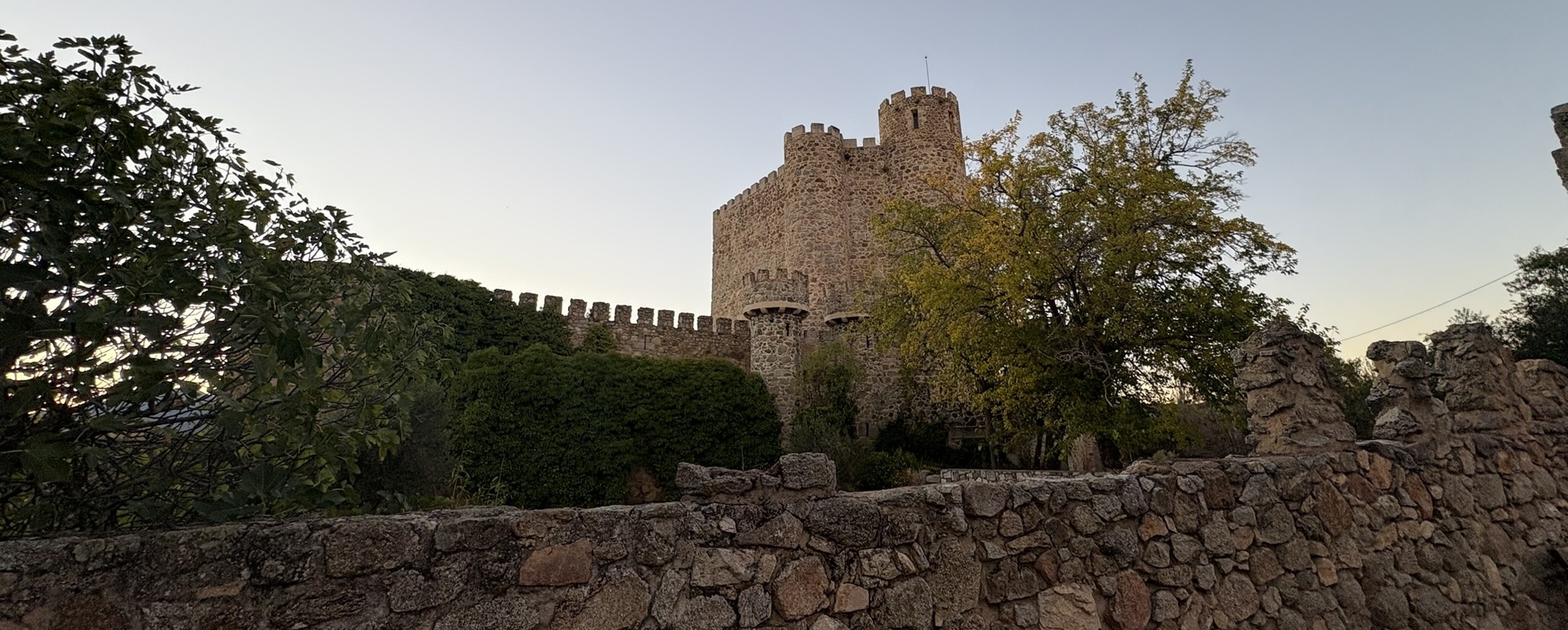 A fortified castle perched on a hilltop at sunset, with warm orange and pink hues in the sky. The medieval structure stands prominently above the town of San Martin de Valdeiglesias.