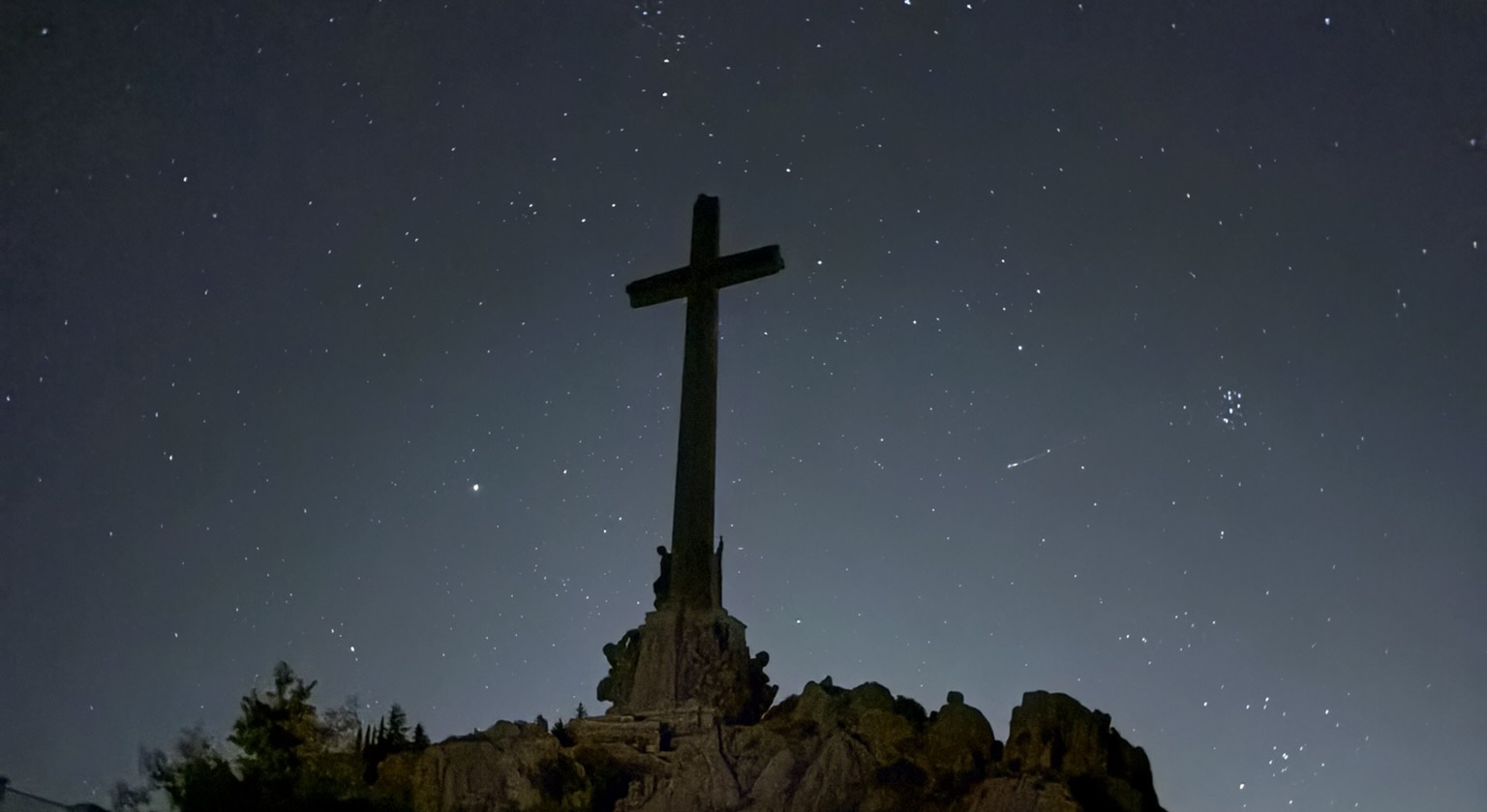 The massive 120-meter cross of Valle de Cuelgamuros illuminated at night against a dark sky, creating a stark and imposing silhouette. The cross dominates the mountainous landscape.