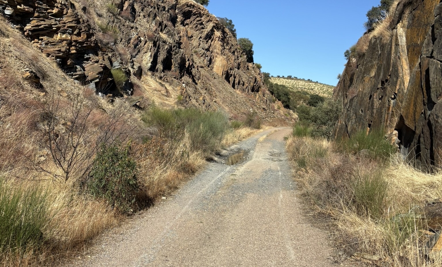 A gravel and broken asphalt path following an old train line through scenic countryside. The Via Verde de la Jara winds through hills with vegetation on both sides.