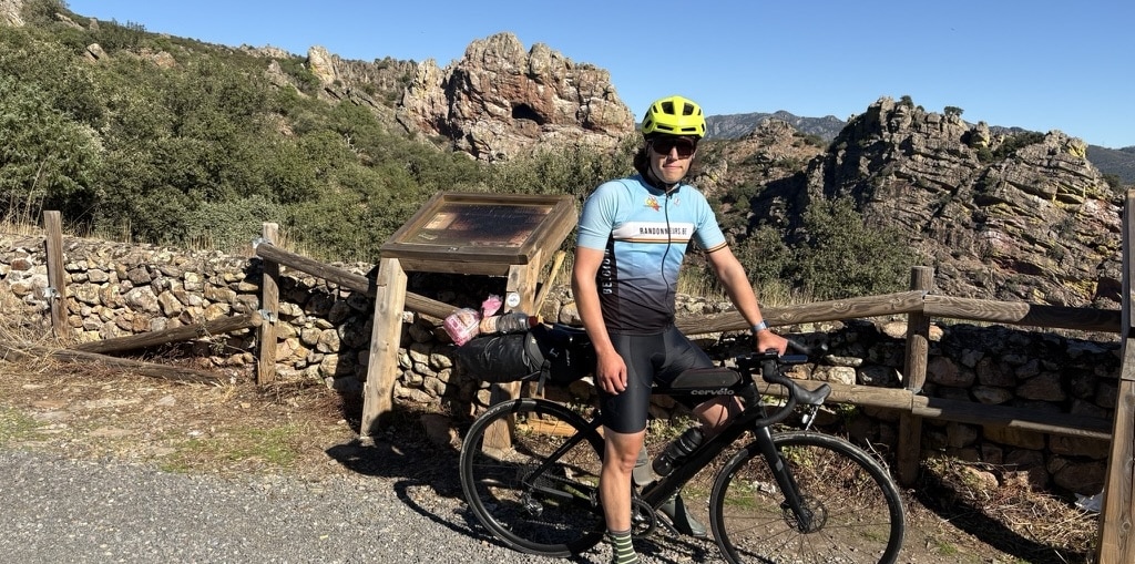 A cyclist with their bike standing at a mountain viewpoint, with dramatic cliffs and valleys of the Sierra de Villuercas in the background under a blue sky.