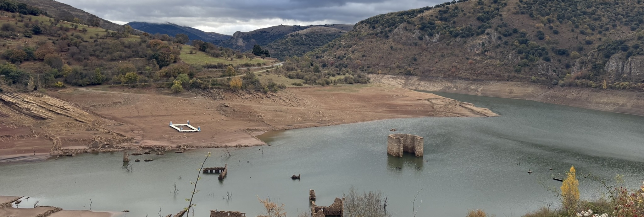 The Embalse de Mansilla reservoir with low water levels revealing the old flooded village of Mansilla de la Sierra. Buildings and structures are visible in the partially drained reservoir surrounded by hills.